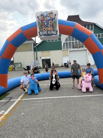 Two families gathered with plush animal scooters under the Come Ride Along arch at a fairground