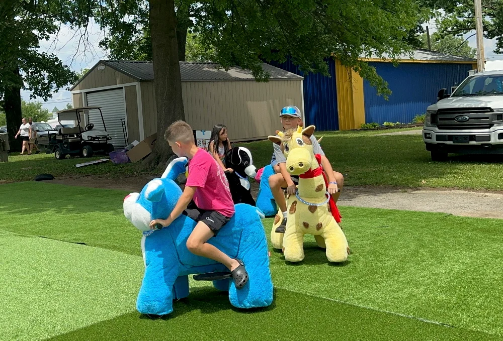 Children riding blue dog and yellow giraffe plush scooters on green turf at an outdoor event