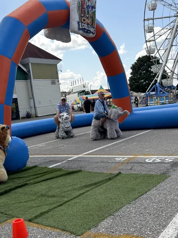 Two riders on plush scooters passing under the inflatable arch at a fair, Ferris wheel behind
