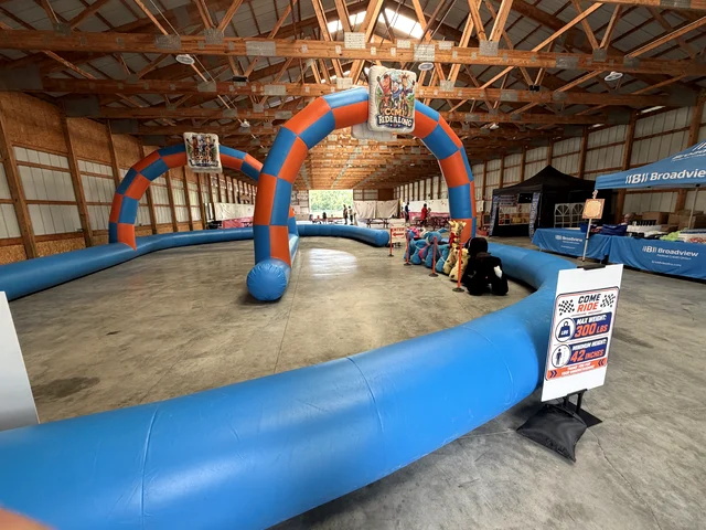 The blue and orange inflatable race track set up inside a barn, with the entry arch and animal scooters parked at the side