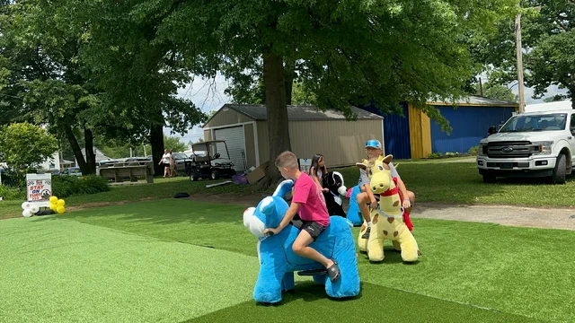 Two children riding plush scooters on the green turf track laid over grass